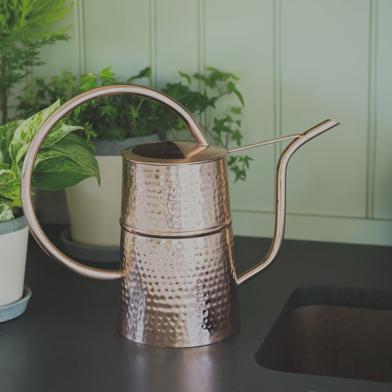 hammered copper watering can sitting on a black countertop next to a sink and houseplants